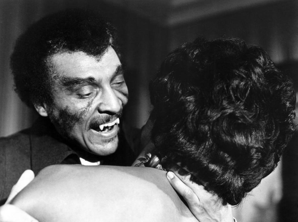 A close-up black and white image of a man with distinctive facial features, smiling and showing his teeth while embracing a woman from behind, whose face is not visible.
