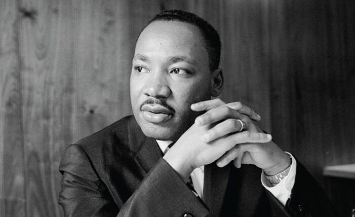 A black and white portrait of a man in a suit, sitting with his hands clasped together, looking thoughtfully to the side.