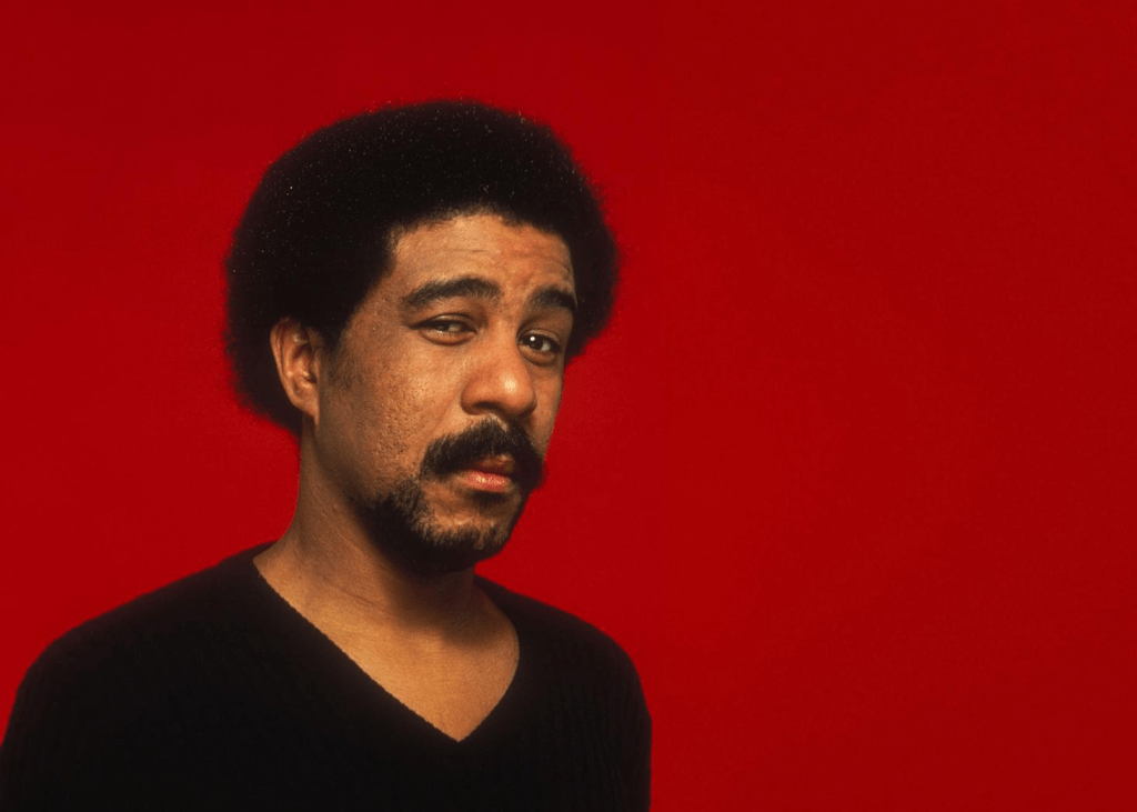 A man with an afro hairstyle is looking slightly sideways against a solid red background, wearing a black sweater.