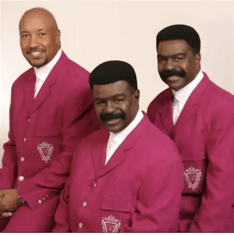 Three men posing together, all wearing matching pink suits with white shirts, against a neutral background.