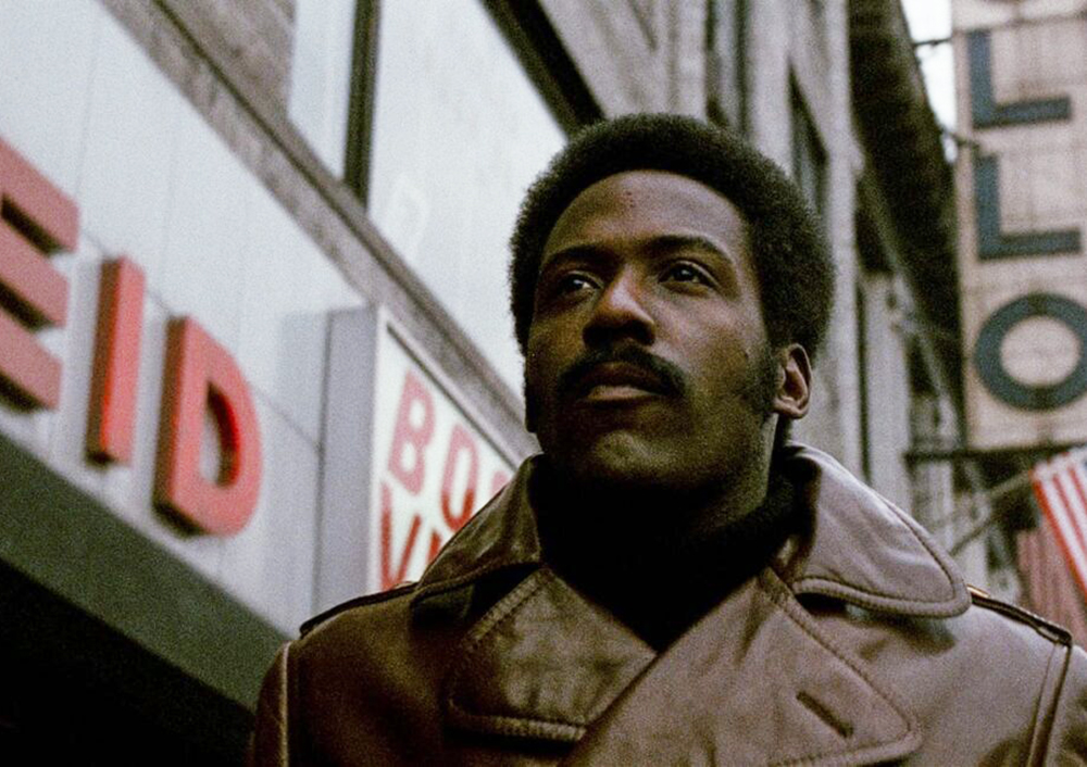 A man with an afro hairstyle, wearing a brown coat, looking upwards in an urban setting with buildings and signs in the background.