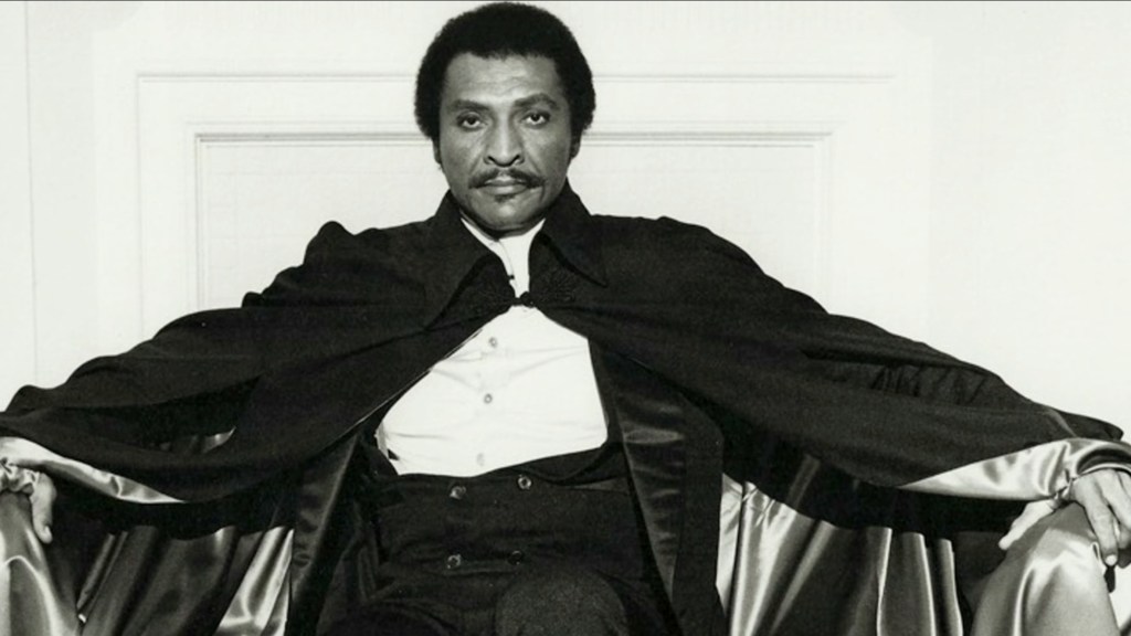 A man seated in a dramatic pose, wearing a black cape over formal attire, exuding an air of sophistication and style.